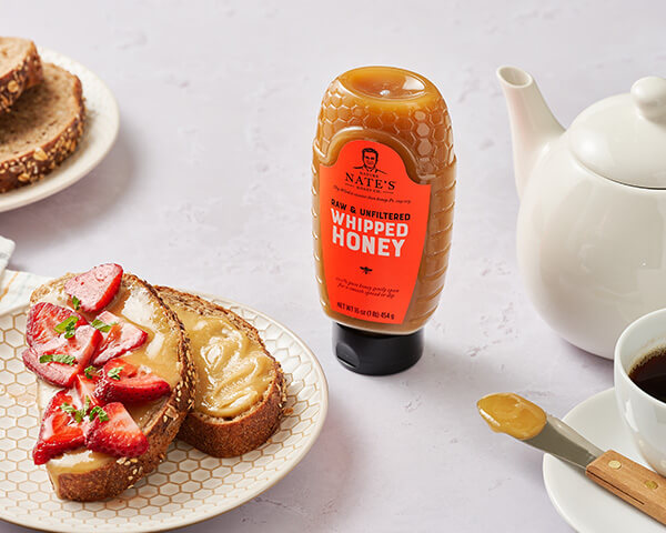 Bottle of Nate’s whipped honey, toast with whipped honey and strawberries, teapot, cozy breakfast setup.