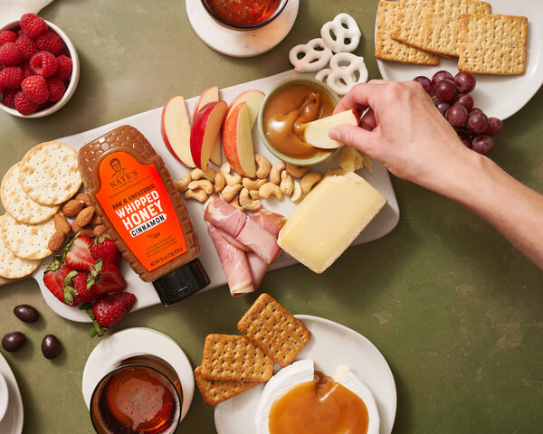 A variety of snacks including cheese, fruit, and crackers, with a bottle of Nate's whipped honey cinnamon.