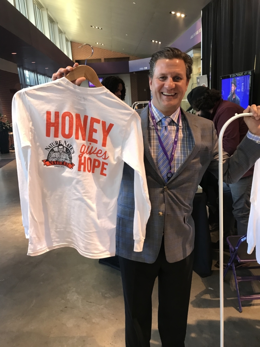man is holding a white t-shirt with the words HONEY gives HOPE, smiling at an event.