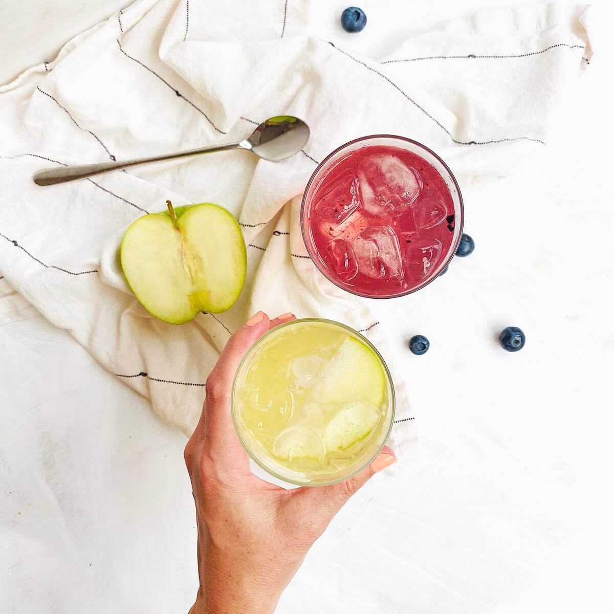 A hand holding a lemon drink with a red drink, apple, berries and spoon on cloth.