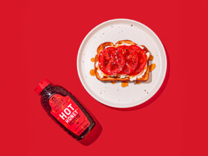A bottle of Nate’s hot honey beside a plate with tomato & ricotta toast on a red background. 