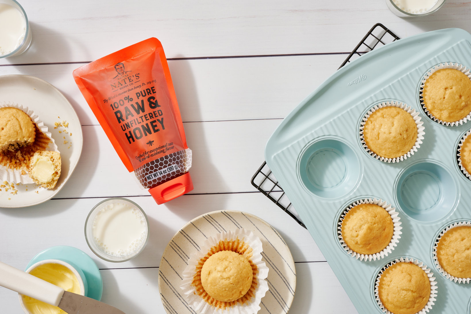Honey muffins on a cooling rack and plates, with a Nate’s honey pouch, butter and milk on a white wooden table.