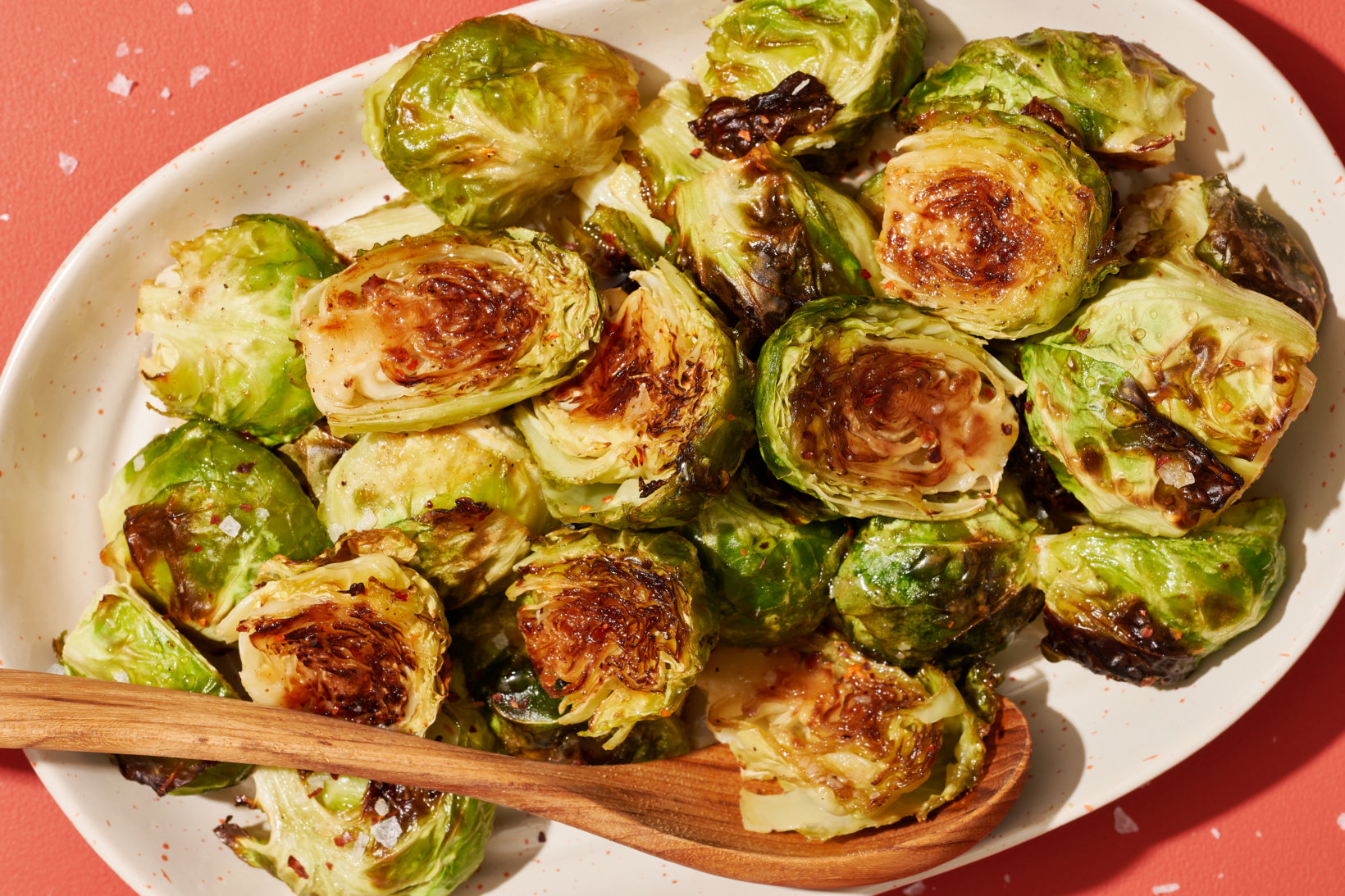 Roasted brussels sprouts glazed with Nate’s hot honey on a plate with a wooden spoon on a red background.