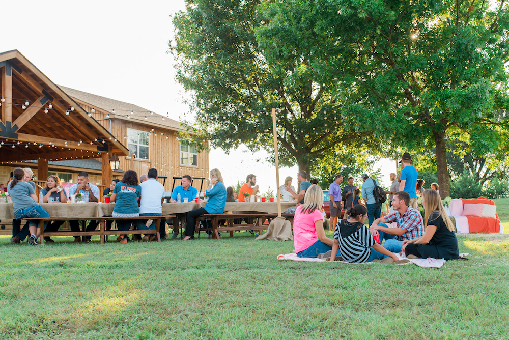 Outdoor gathering with people at picnic tables on a lawn.