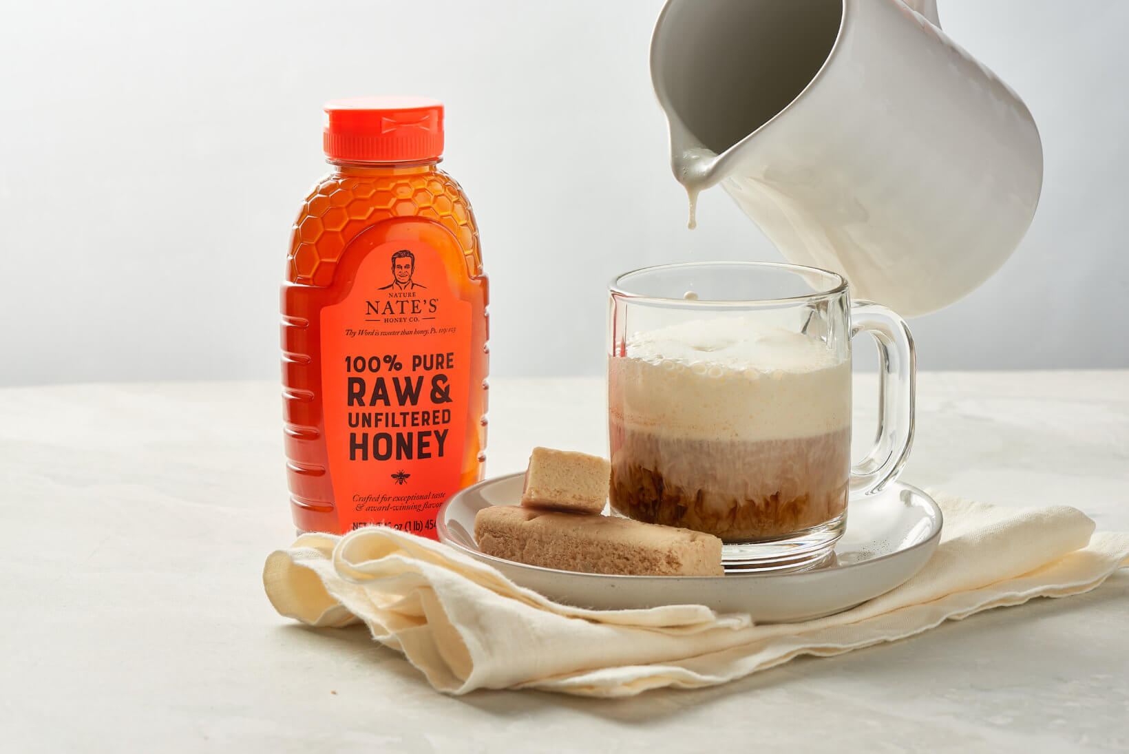 Latte being poured into a glass mug with frothed milk, shortbread biscuits  and a bottle of Nate’s honey on a cloth.