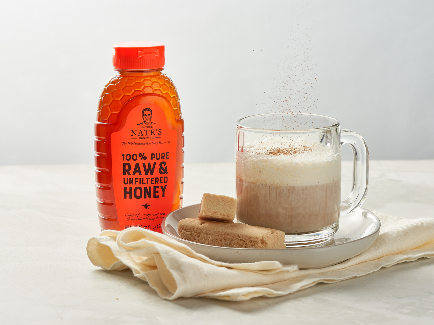 A bottle of Nate’s honey next to a steaming mug of latte accompanied by shortbread cookies on a white background.