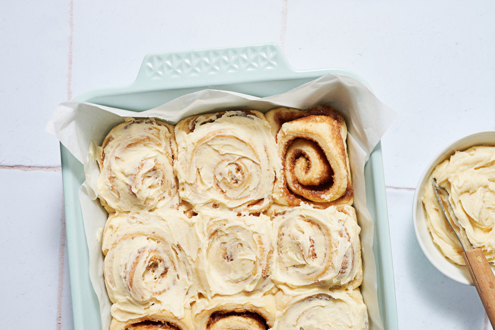 Honey cinnamon rolls with cream frosting in a baking dish, one roll unfrosted, on a white countertop.