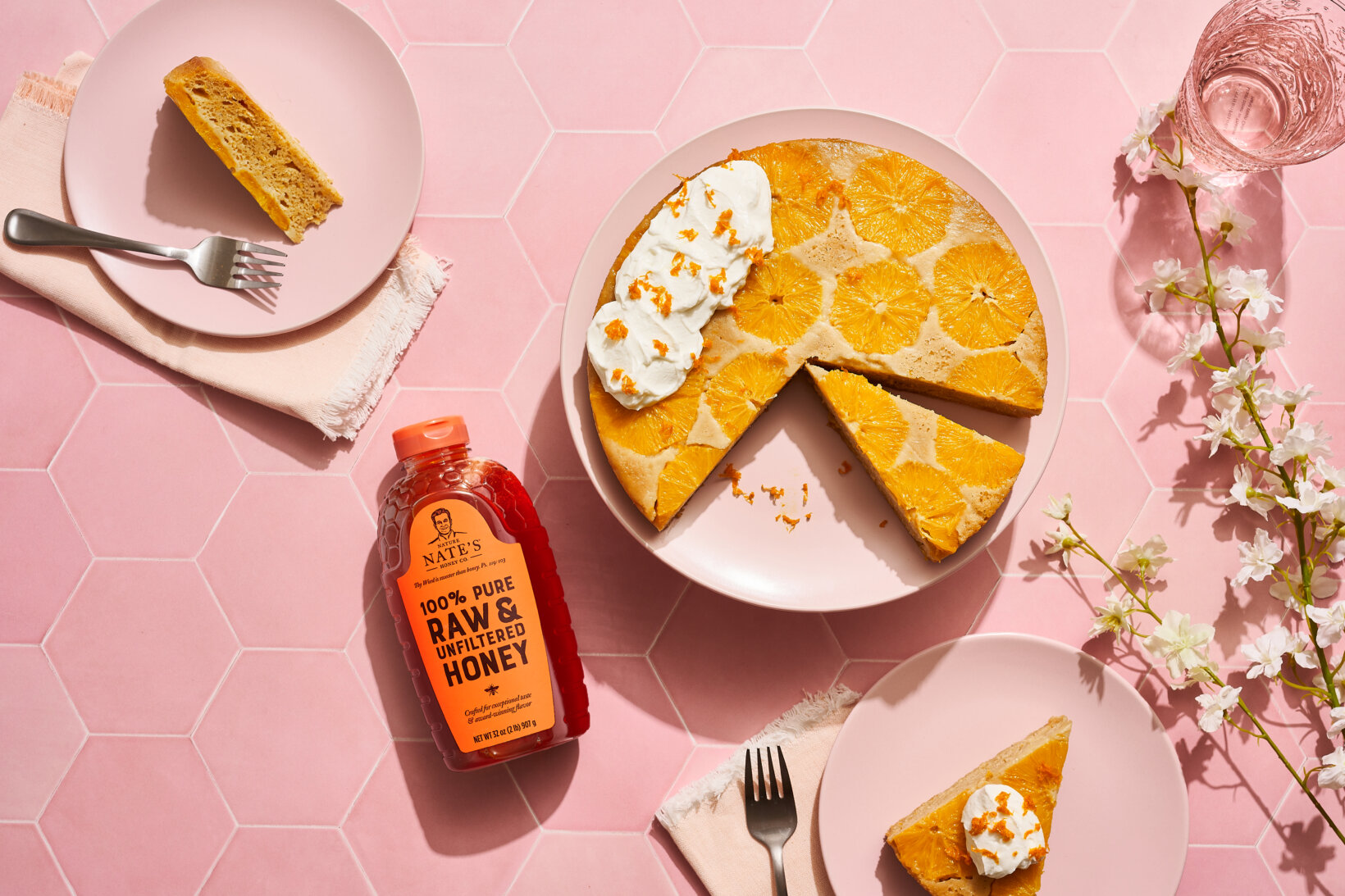 Olive oil cake sliced and garnished with cream next to a bottle of Nate’s honey on a pink table.