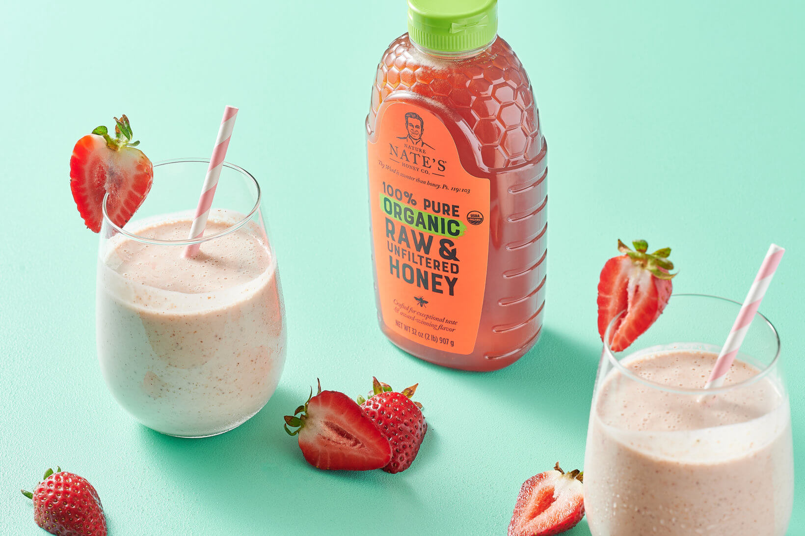 Strawberry smoothies with Nate’s organic honey bottle with a green cap and fresh strawberries on a teal background.
