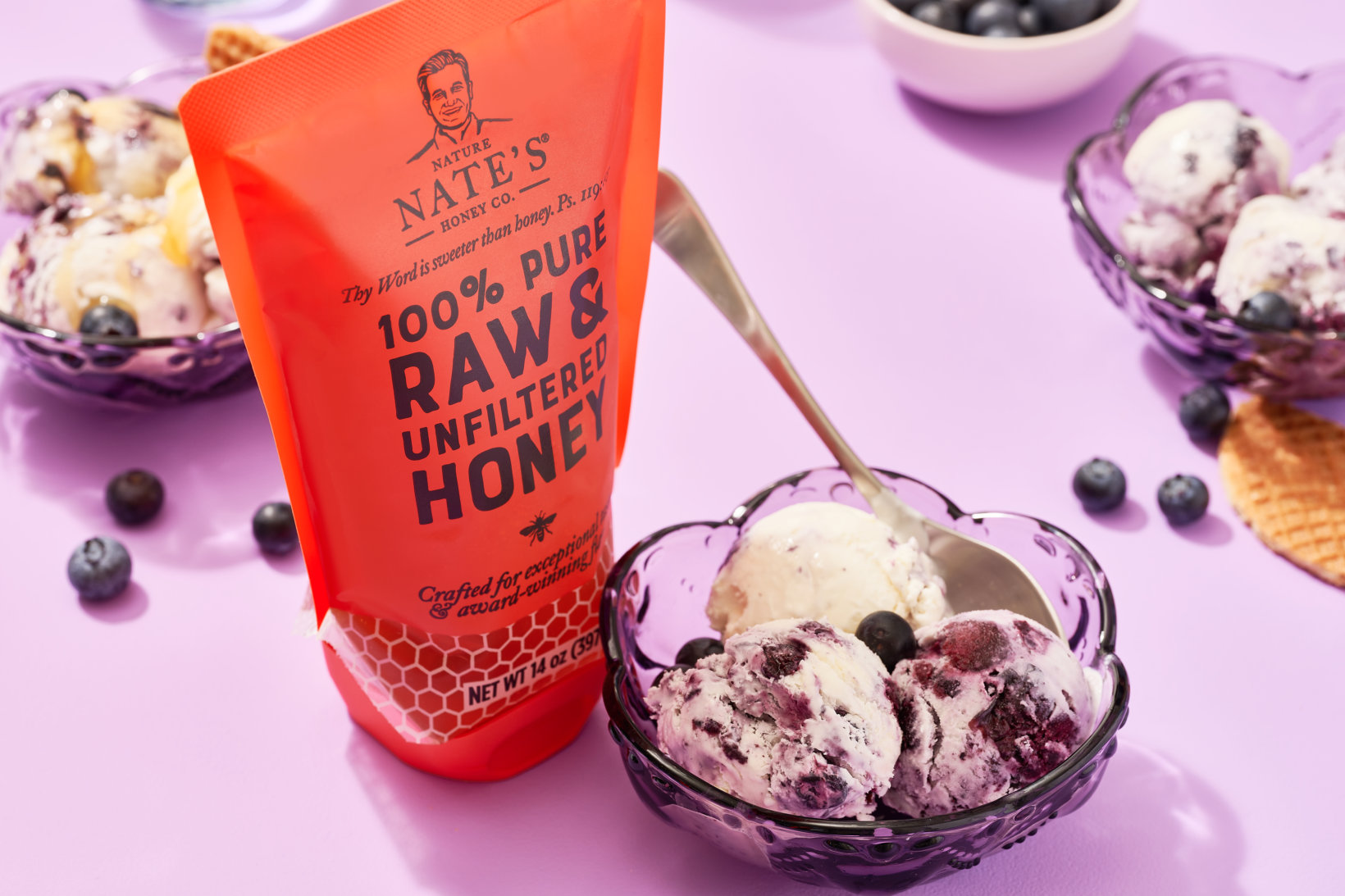 A pouch of Nate's 100% pure raw & unfiltered honey beside a bowl of blueberry ice cream on a purple background.