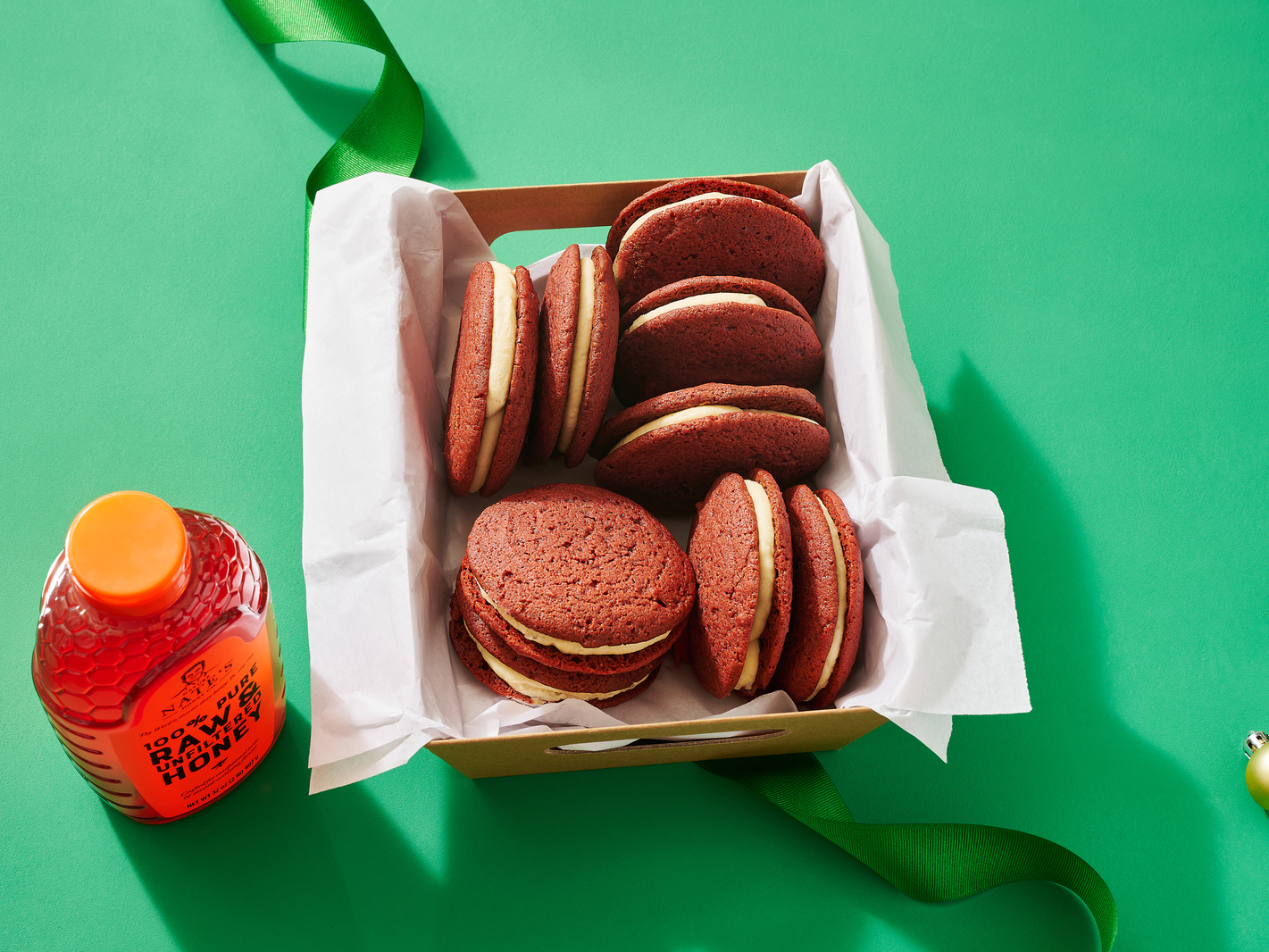 Red velvet whoopie pies in a box with a bottle of Nate’s honey on a green background.