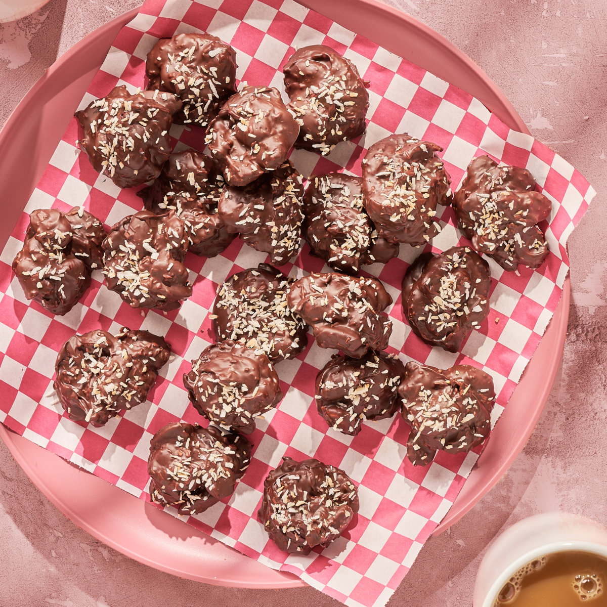 Chocolate almond clusters made with honey and sprinkled with coconut on checked paper in a pink plate and cup of coffee.