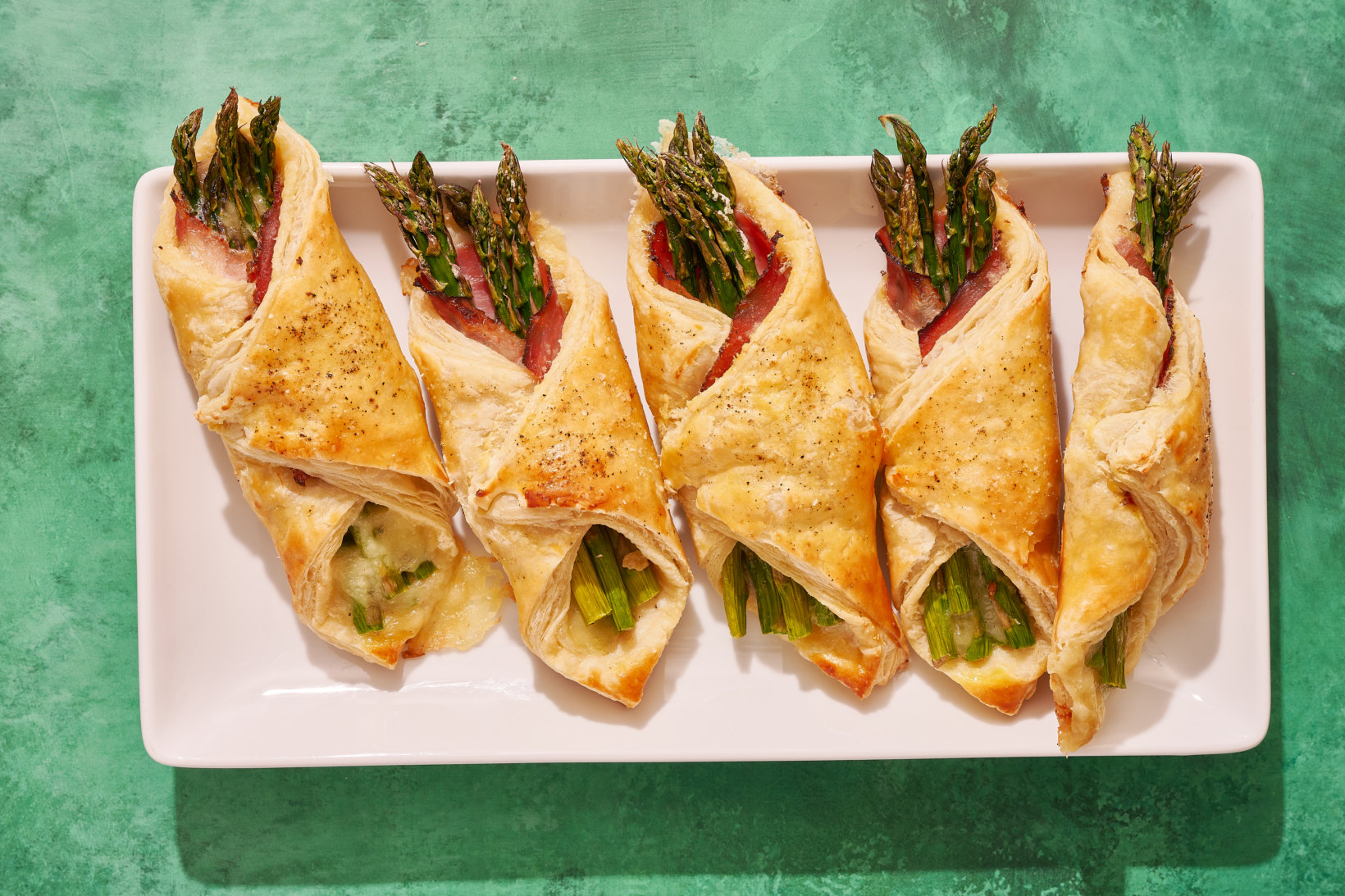 Asparagus, ham and honey wrapped in a puff pastry arranged on a rectangular plate against a textured green background.