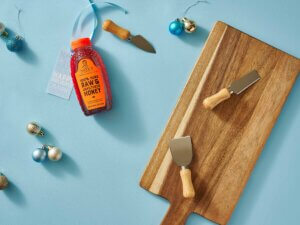 A bottle of Nates honey with a gift tag, cheese knives and a wooden board amidst scattered Christmas ornaments.