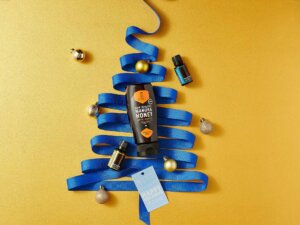 Nate’s manuka honey bottle laying on a blue ribbon shaped like a tree with ornaments on yellow background.