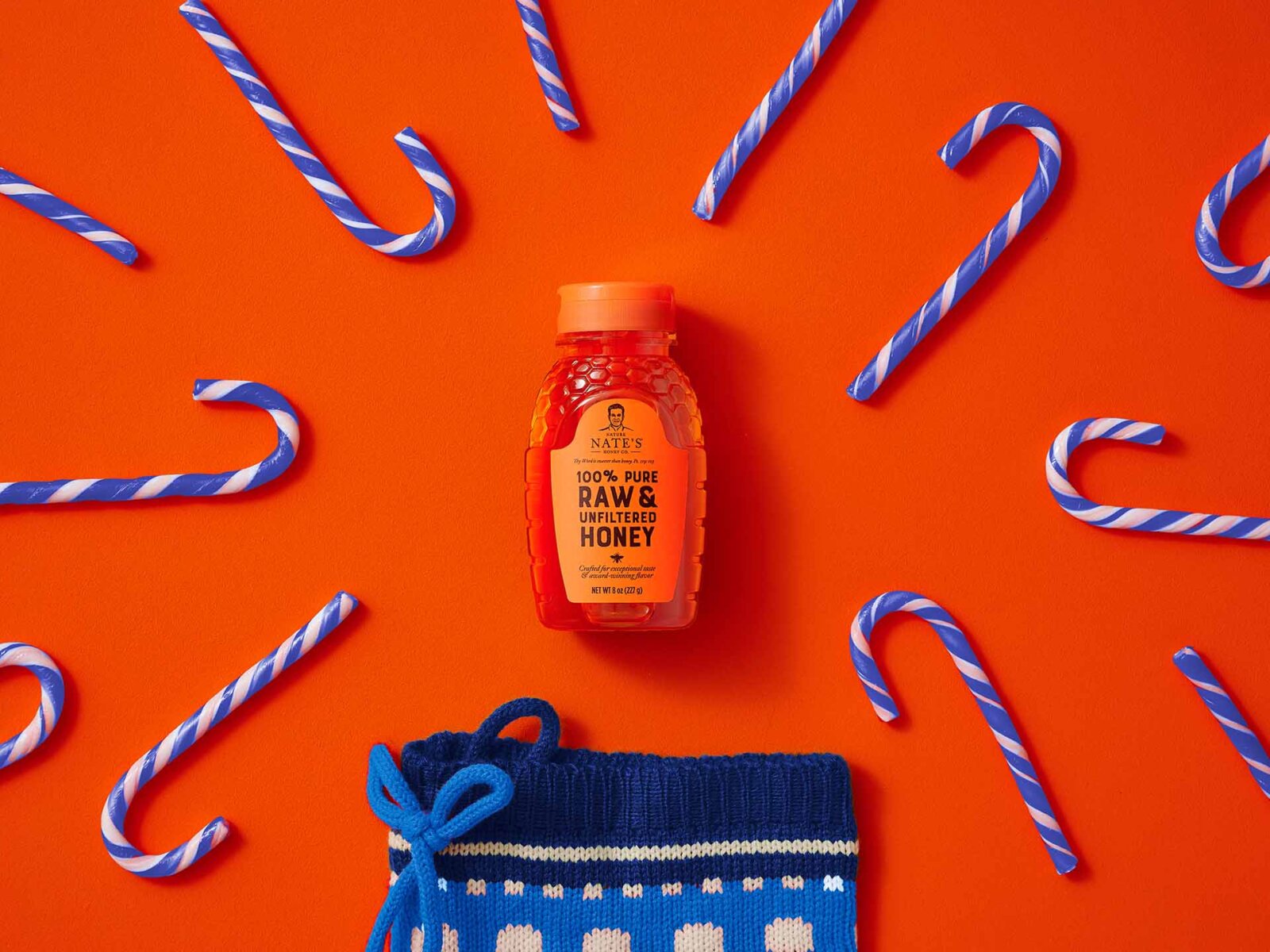 A bottle of Nate’s honey on an orange background with blue candy canes and a blue stocking.