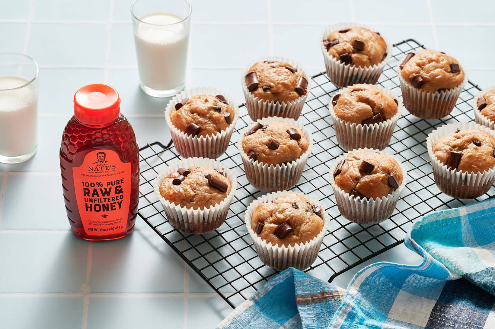 Muffins on a cooling rack, a bottle of Nate's honey and glasses of milk  on a tiled counter with a blue checkered towel.