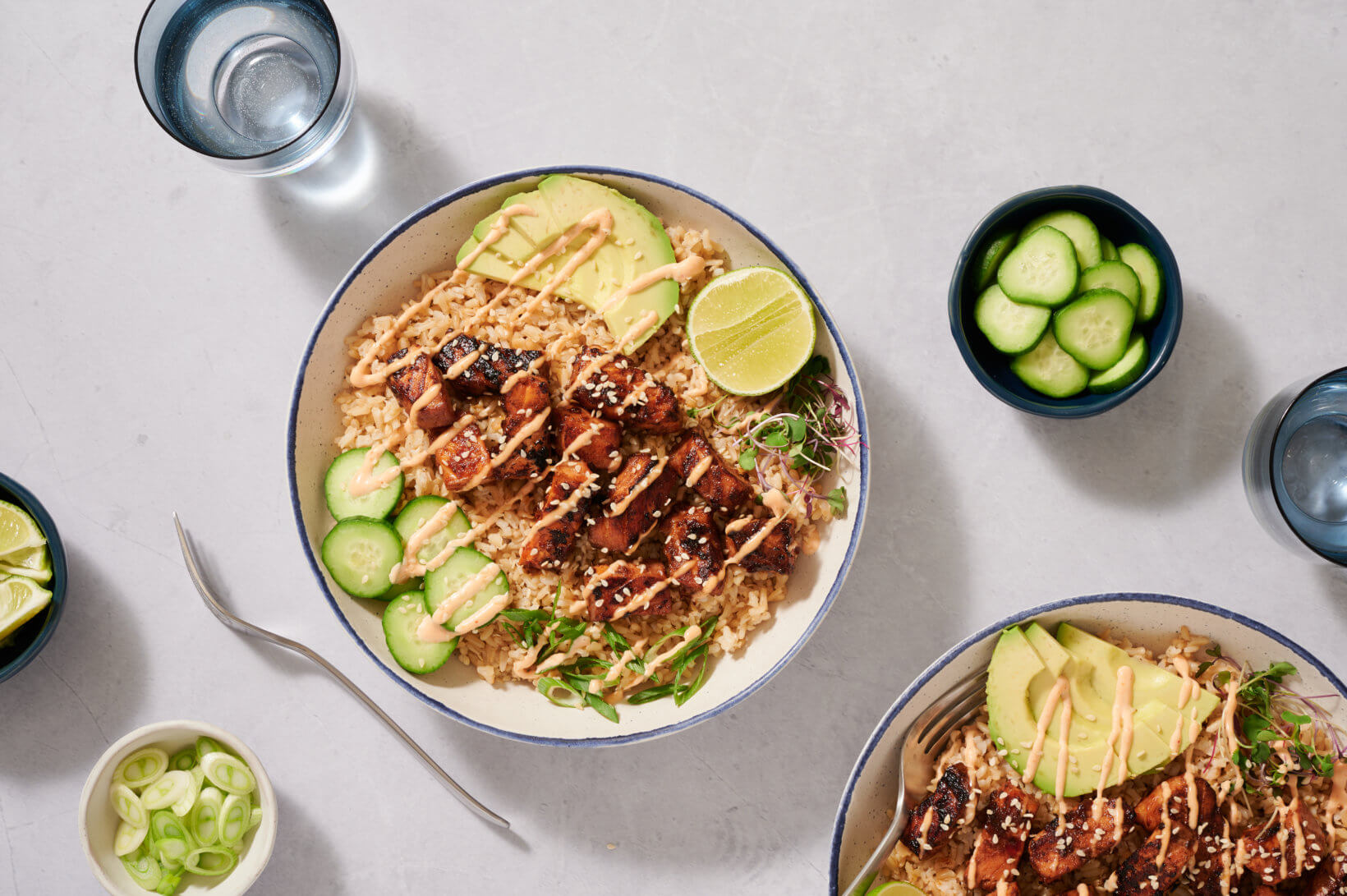 Bowls of rice topped with grilled chicken, avocado, cucumber, lime, and drizzled sauce, accompanied by glasses of water and small bowls of sliced cucumber and green onions.