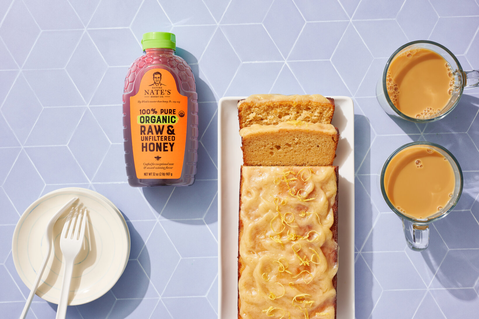 Sliced lemon loaf cake, a bottle of Nate’s organic honey, two cups of coffee and plates on a blue tile background.