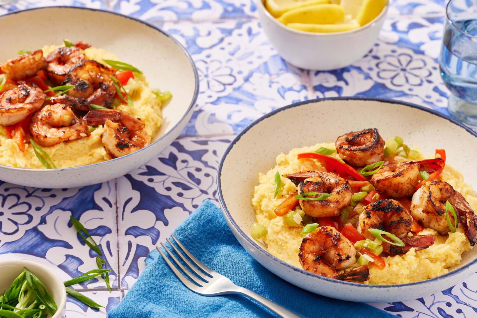Two bowls of shrimp and grits made with hot honey with sliced peppers and green onions on a blue patterned table.