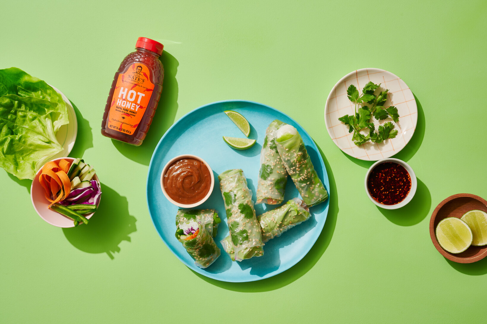 Spring rolls on a plate with a bottle of Nate’s hot honey, dipping sauces and fresh veggies on a green background.