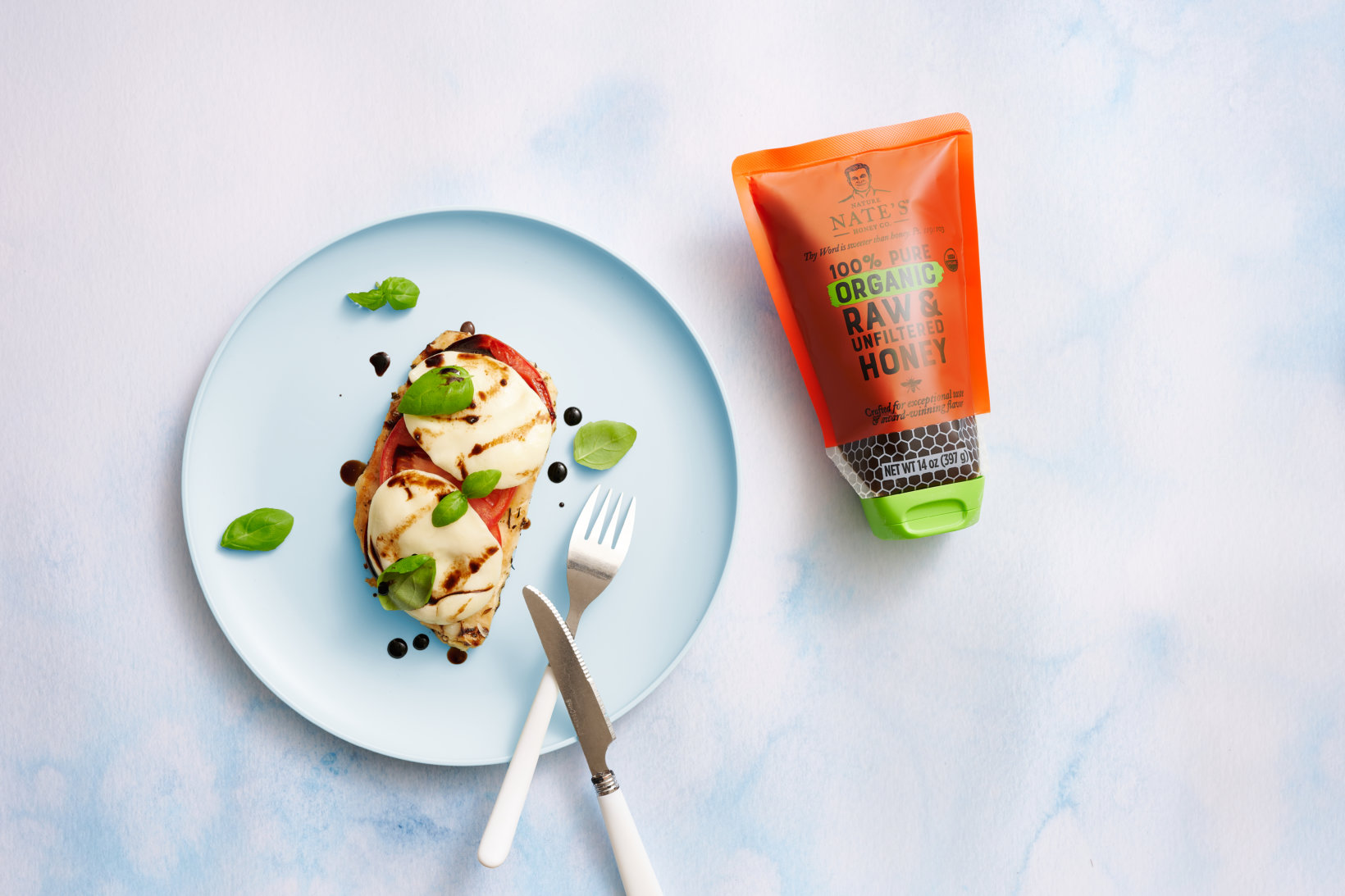 A dish with chicken, mozzarella, tomato and balsamic glaze on a blue plate, a Nate's organic honey pouch, with cutlery.