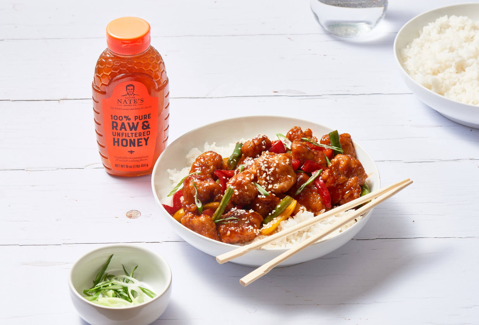 A bottle of Nate’s honey next to a plate of sesame chicken with vegetables over rice, suggesting a honey-infused recipe.