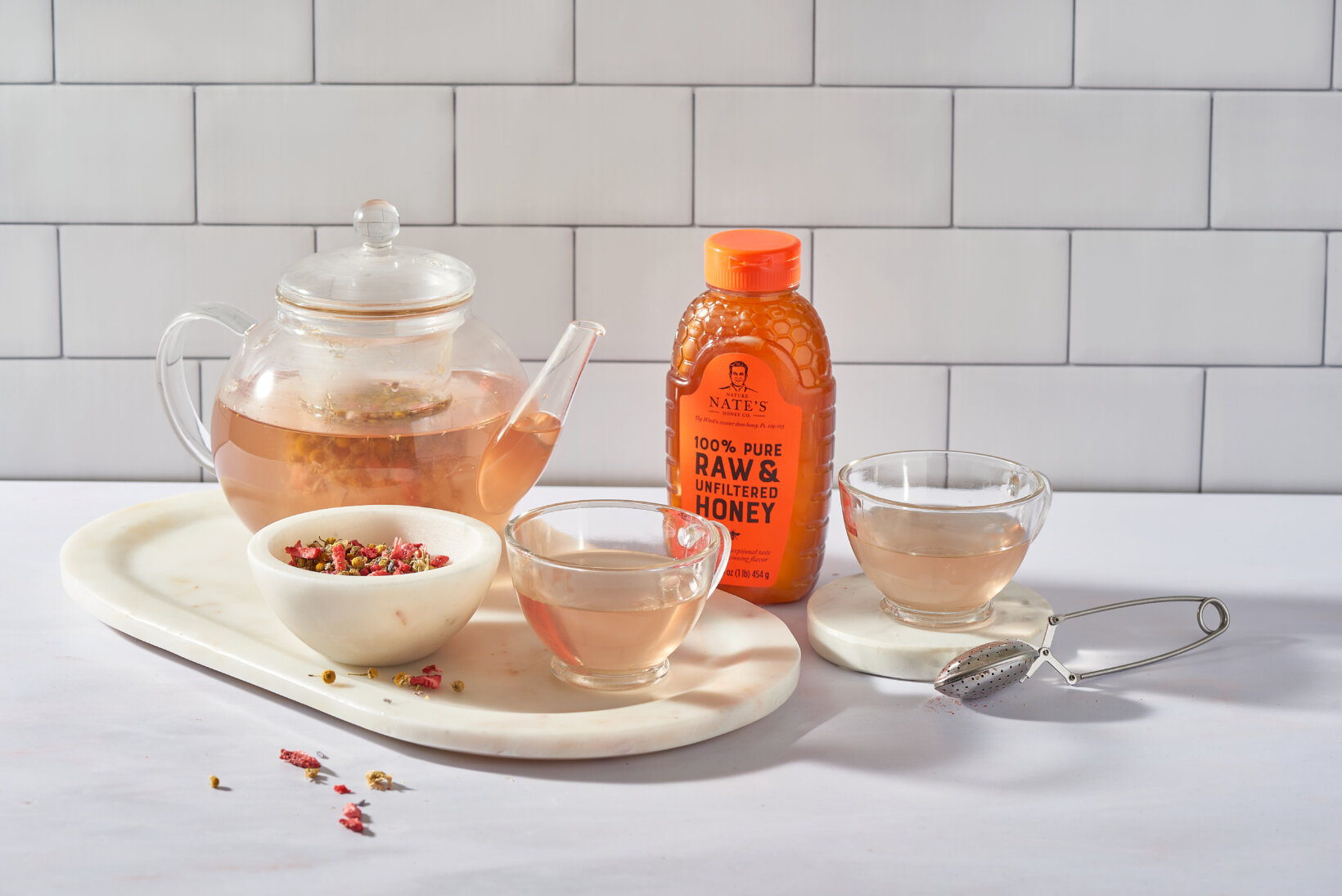 Tea set with clear teapot, two cups, honey bottle, and infuser on a white marble table and subway tile background.