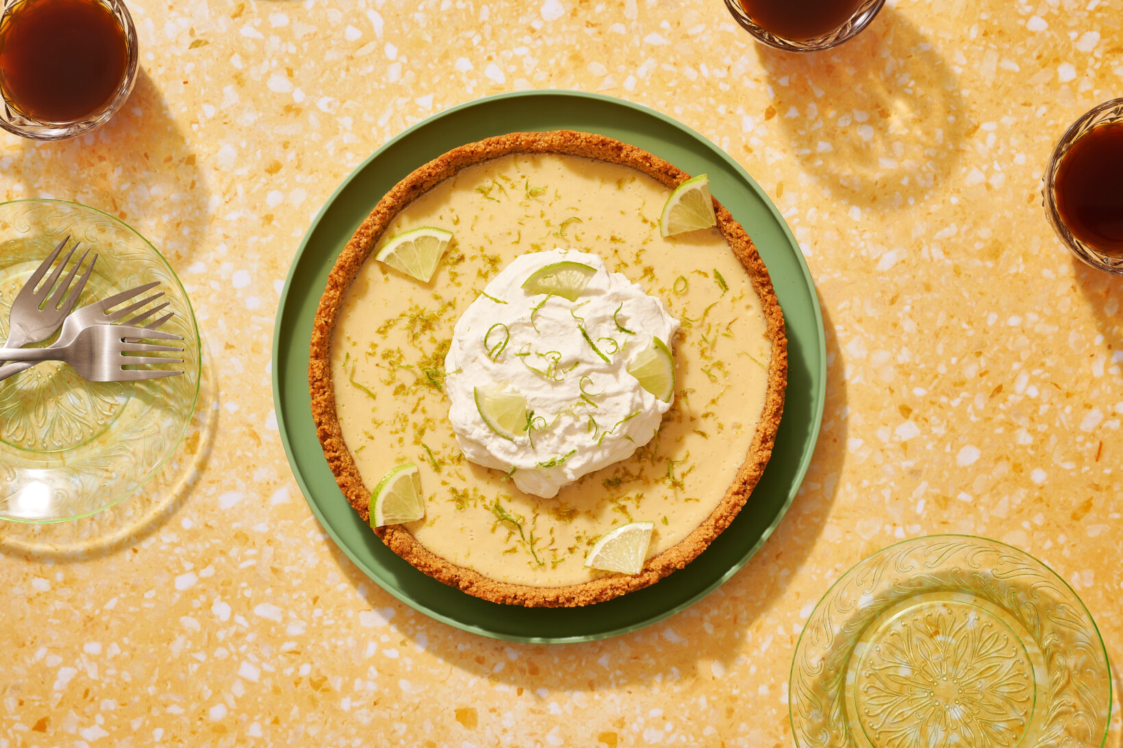 Key lime pie made with Nate’s honey and topped with whipped cream and lime zest on a table with drinks and forks.