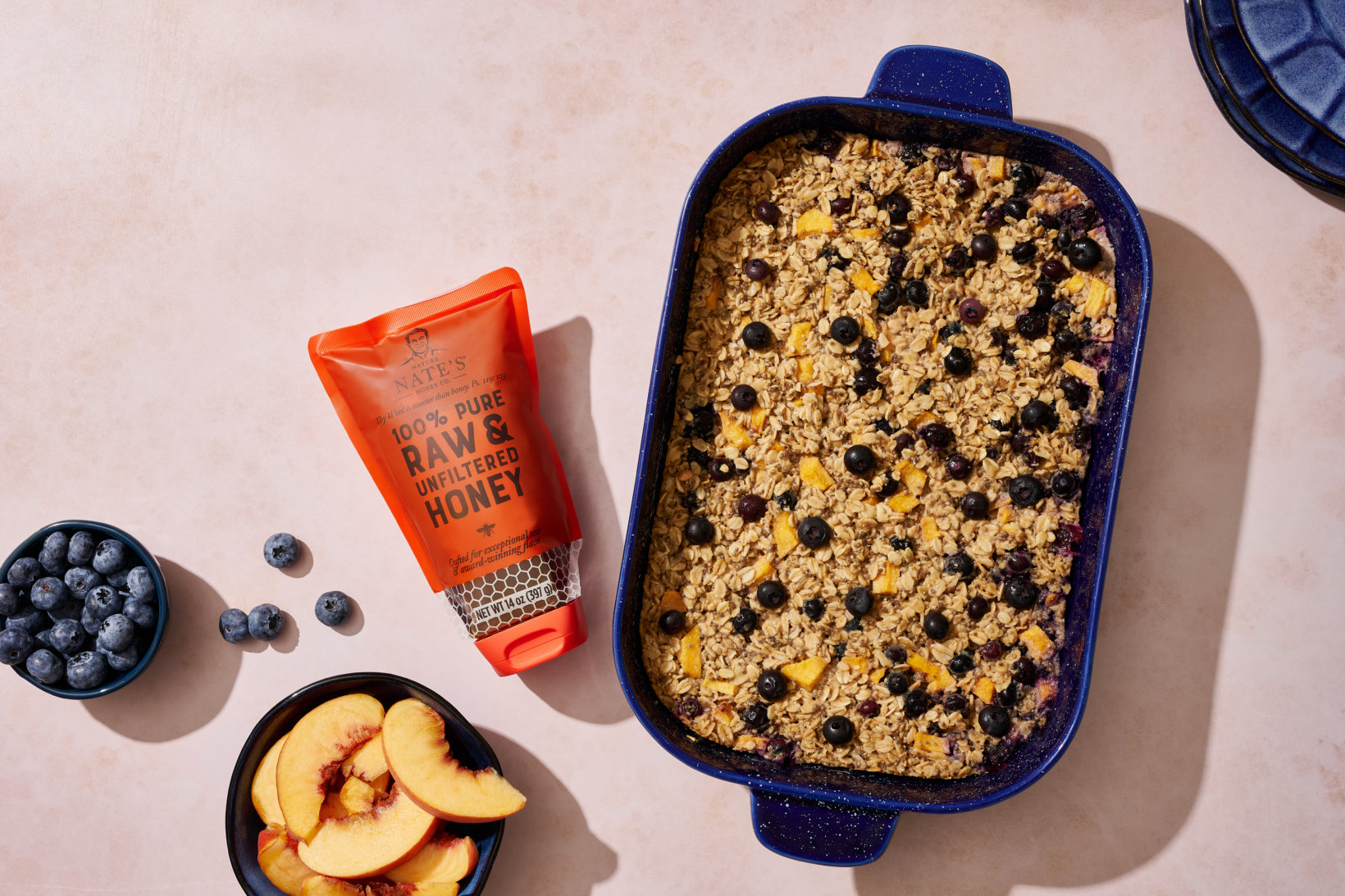 A baked oatmeal dish in a blue dish, a tube of honey, with bowls of blueberries and peach slices on a beige surface.