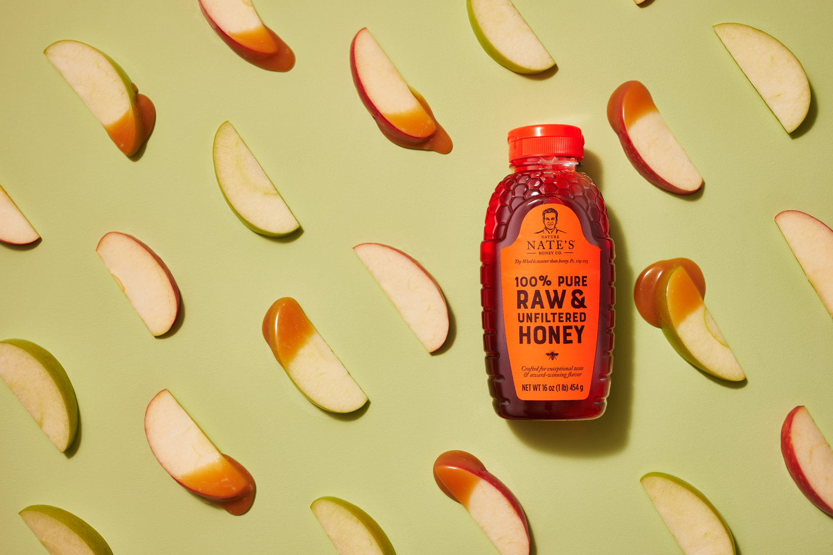 A bottle of Nate's honey with an orange cap surrounded by sliced apple pieces dipped in honey on a green background.