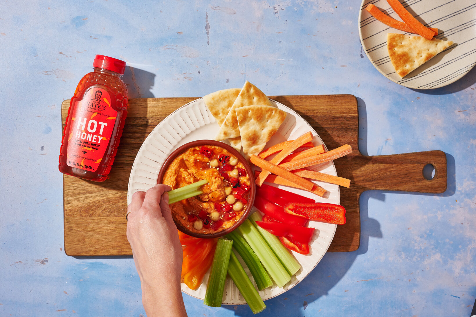 A hand dipping a celery stick into red pepper hummus, with a bottle of Nature Nate's Hot Honey in the background.