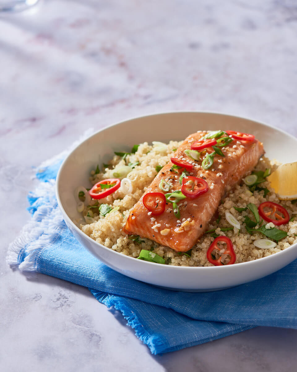 Honey-glazed salmon on couscous with chili slices, herbs in a bowl on a blue napkin.