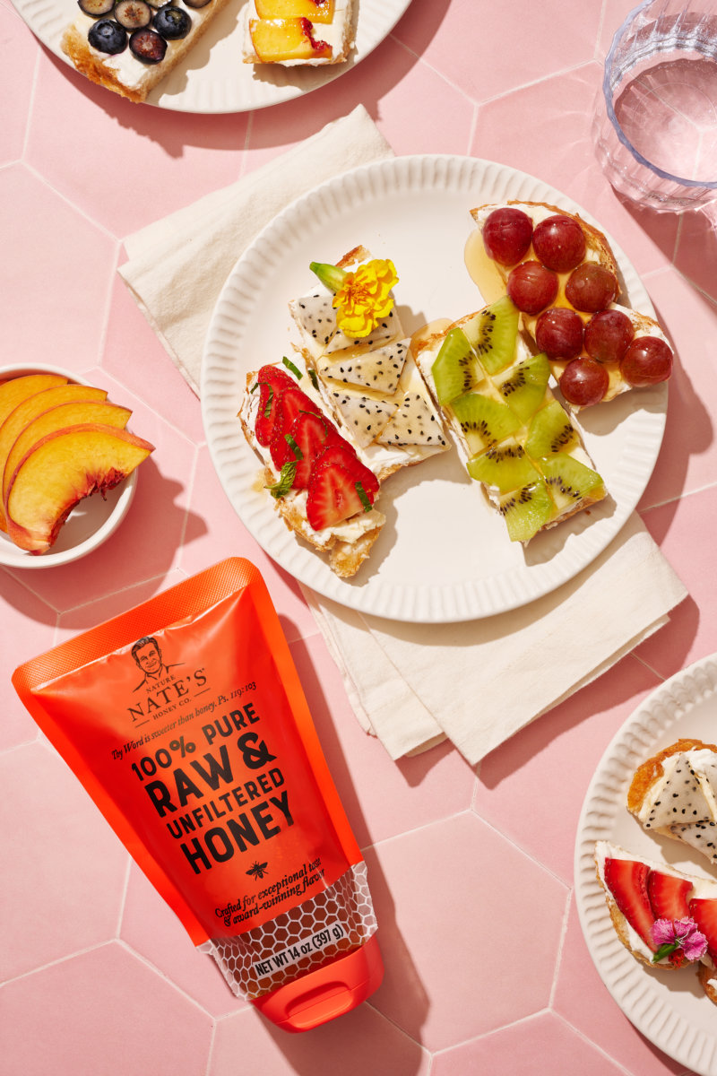 Open-faced fruit toasts on plates with a pouch of Nate’s raw & unfiltered honey, against a pink background.