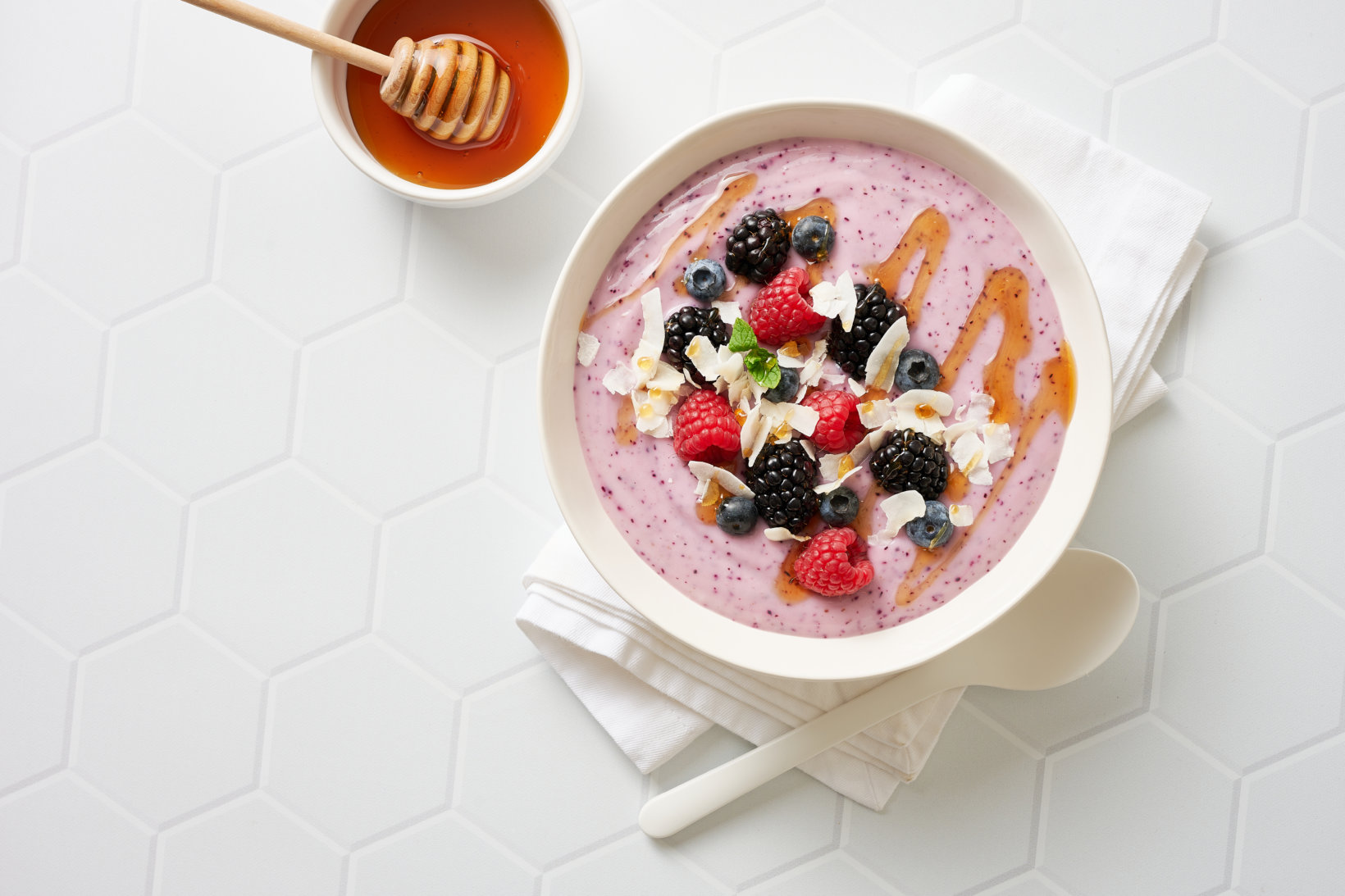 A smoothie bowl with mixed berries and a drizzle of honey, next to a honey dipper on a white hexagonal tile surface.