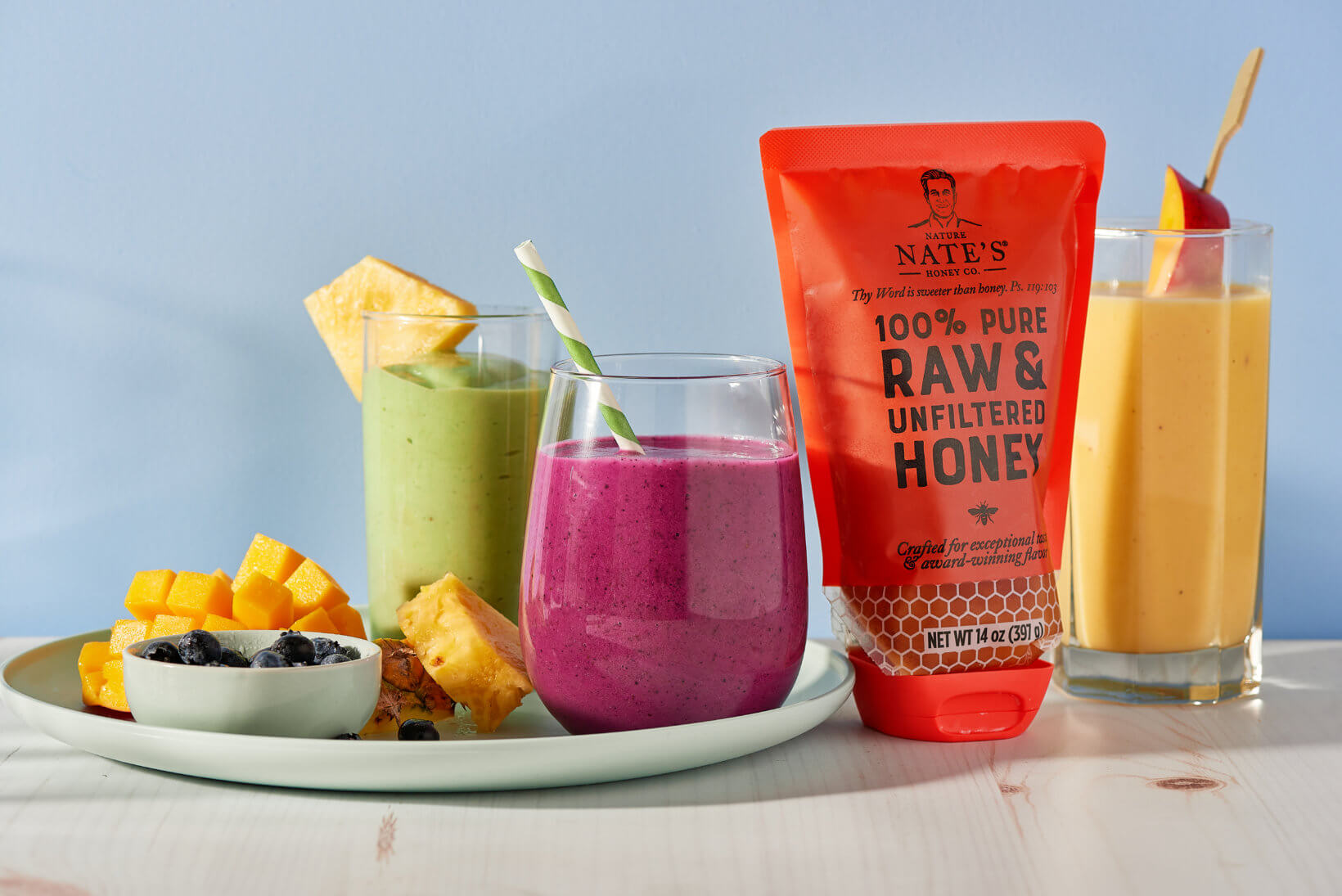 Colorful smoothies in glasses, a plate of fruit and a pouch of Nate’s honey against a blue background.