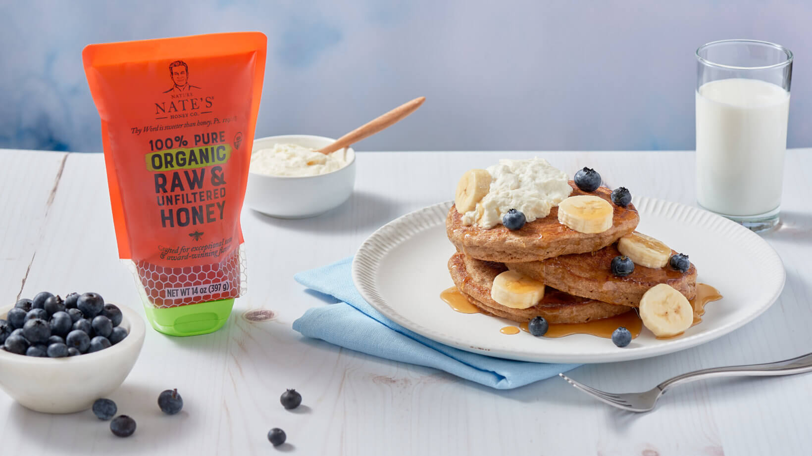 Pancakes with blueberries, bananas and cream beside a Nate’s organic honey pouch, blueberries and a glass of milk.