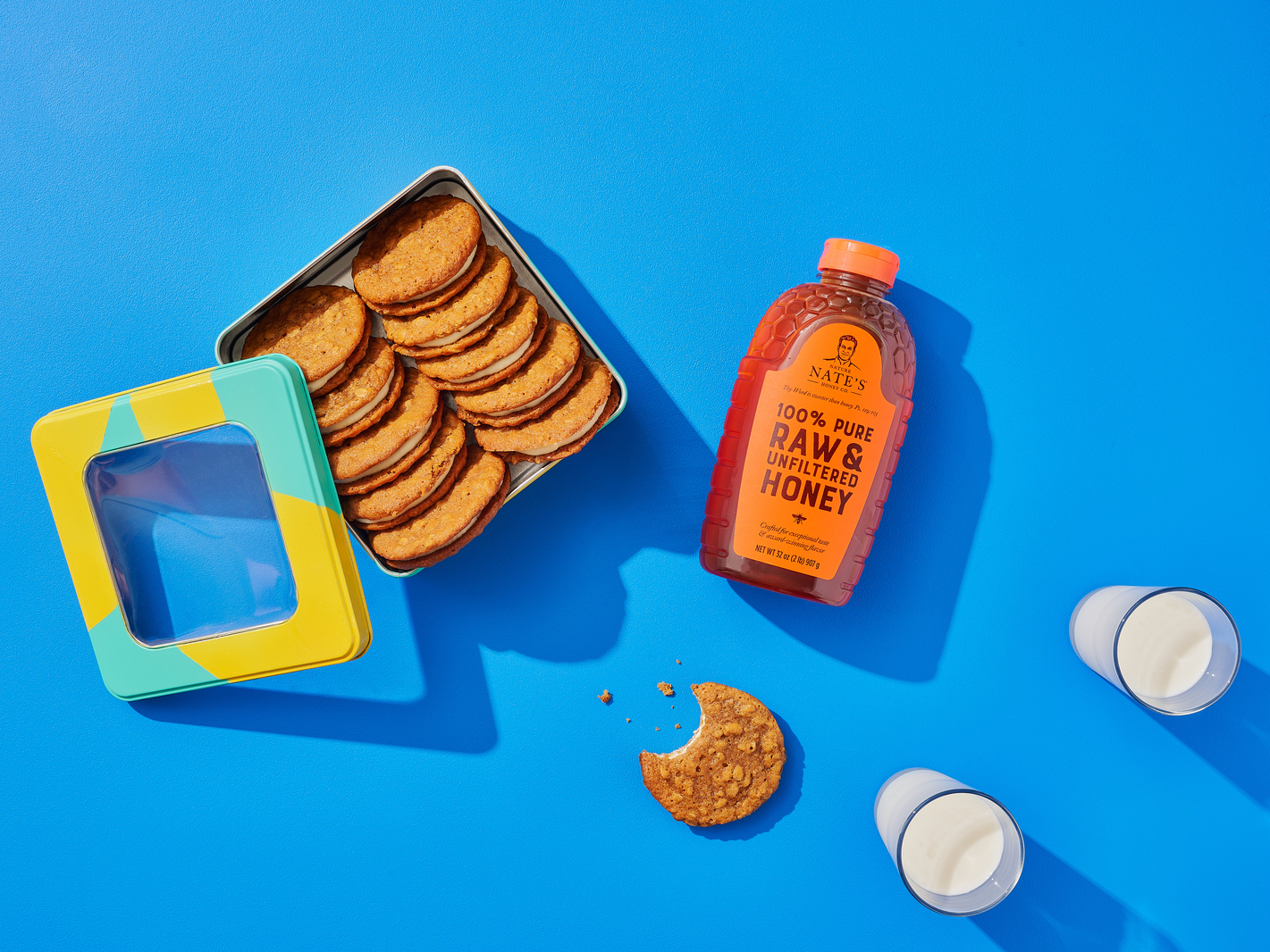 A tin of oatmeal crème pies next to a bottle of Nate’s honey, an oatmeal pie missing a bite and two glasses of milk.