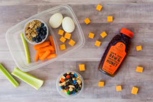 A well-arranged snack setup with boiled eggs, carrots, celery, granola, cheese and a bottle of Nate's raw honey.