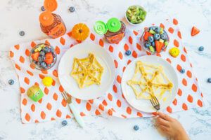 A playful kids’ meal with web-shaped pancakes, fruit cups and two bottles of Nate’s honey, on a patterned tablecloth.