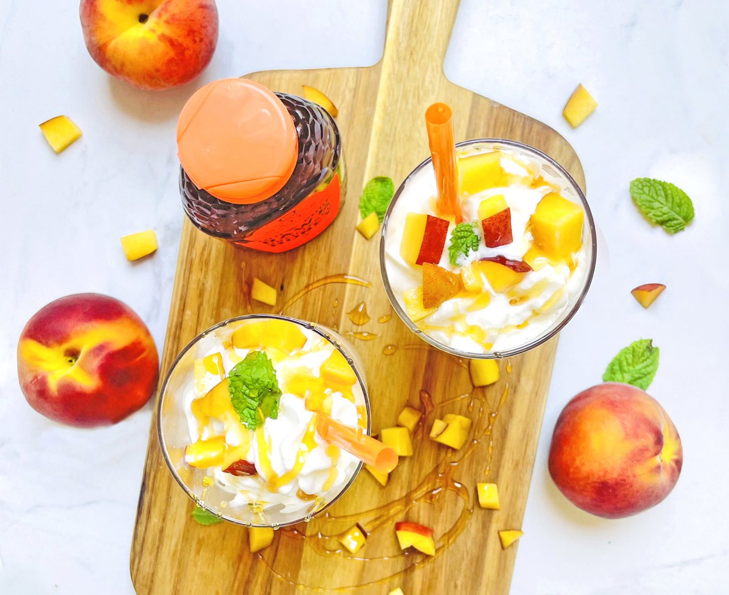 Peach parfaits with mint on a cutting board, Nate's Honey, and fresh peaches on a marble background.