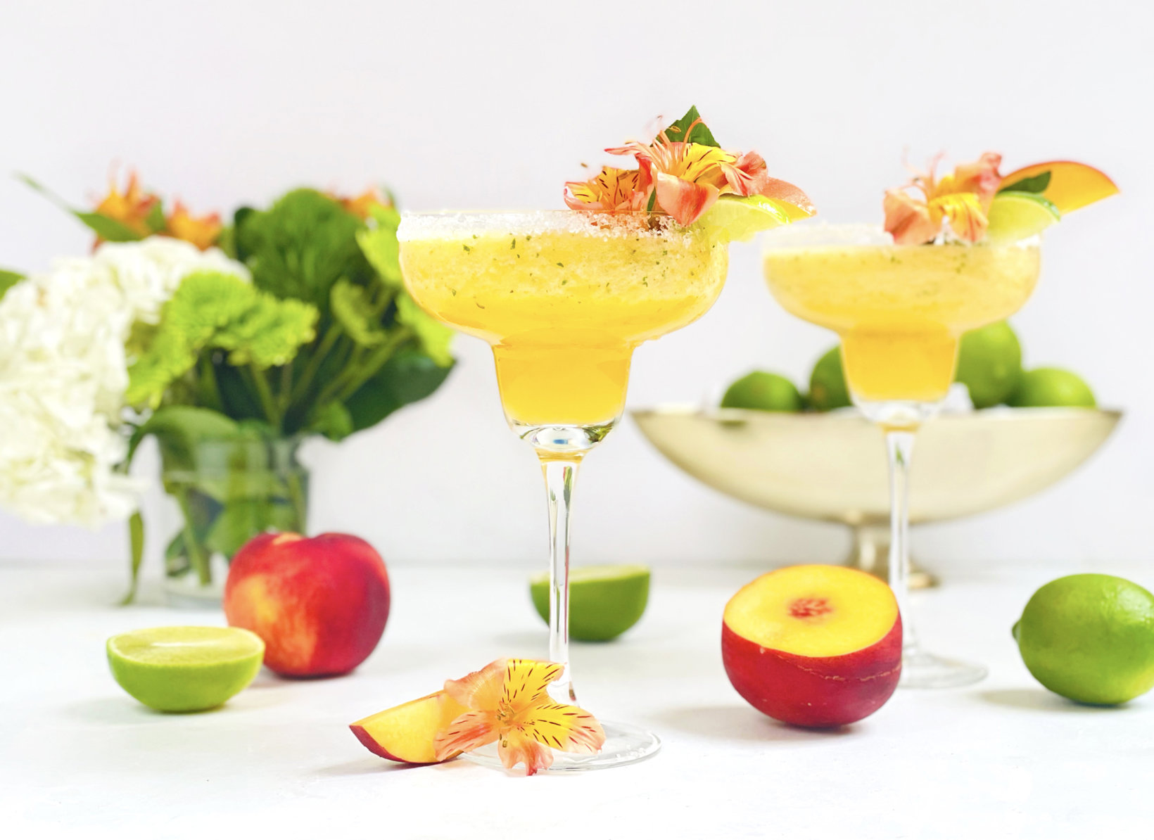 Fruity cocktails in margarita glasses garnished with flowers, with fresh fruit and flowers in the background.