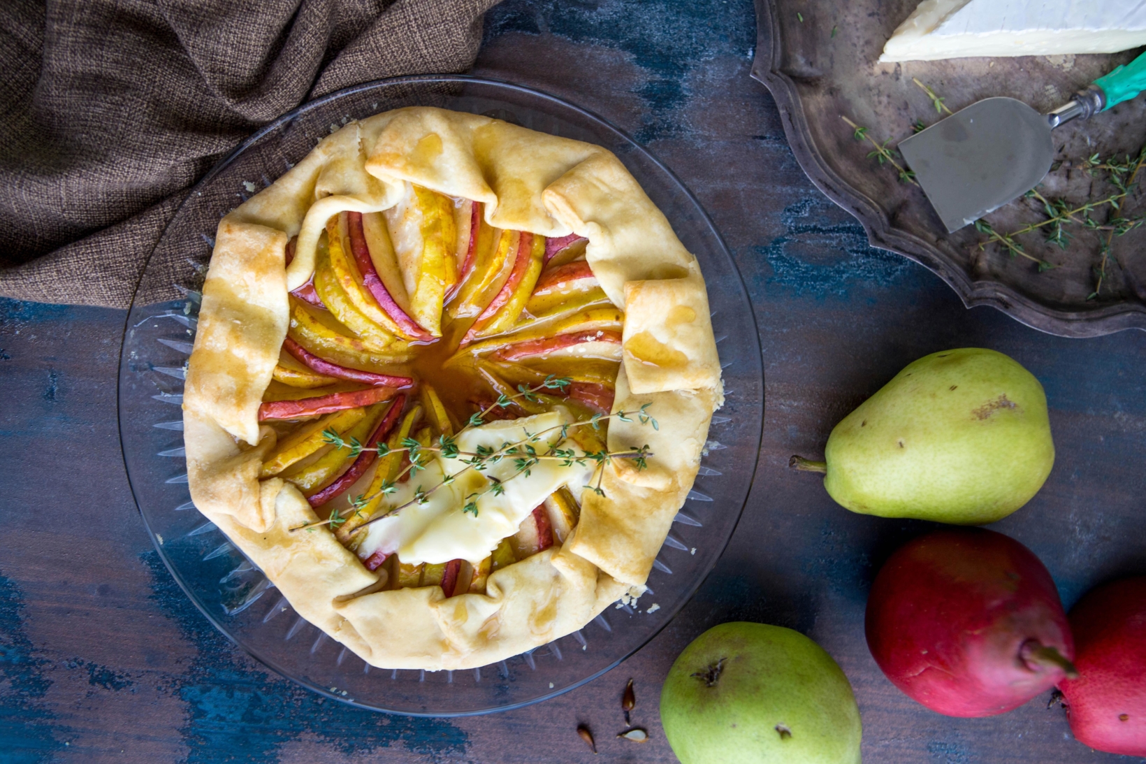 Rustic pear tart topped with brie cheese, on a blue wooden table with fresh fruits around.