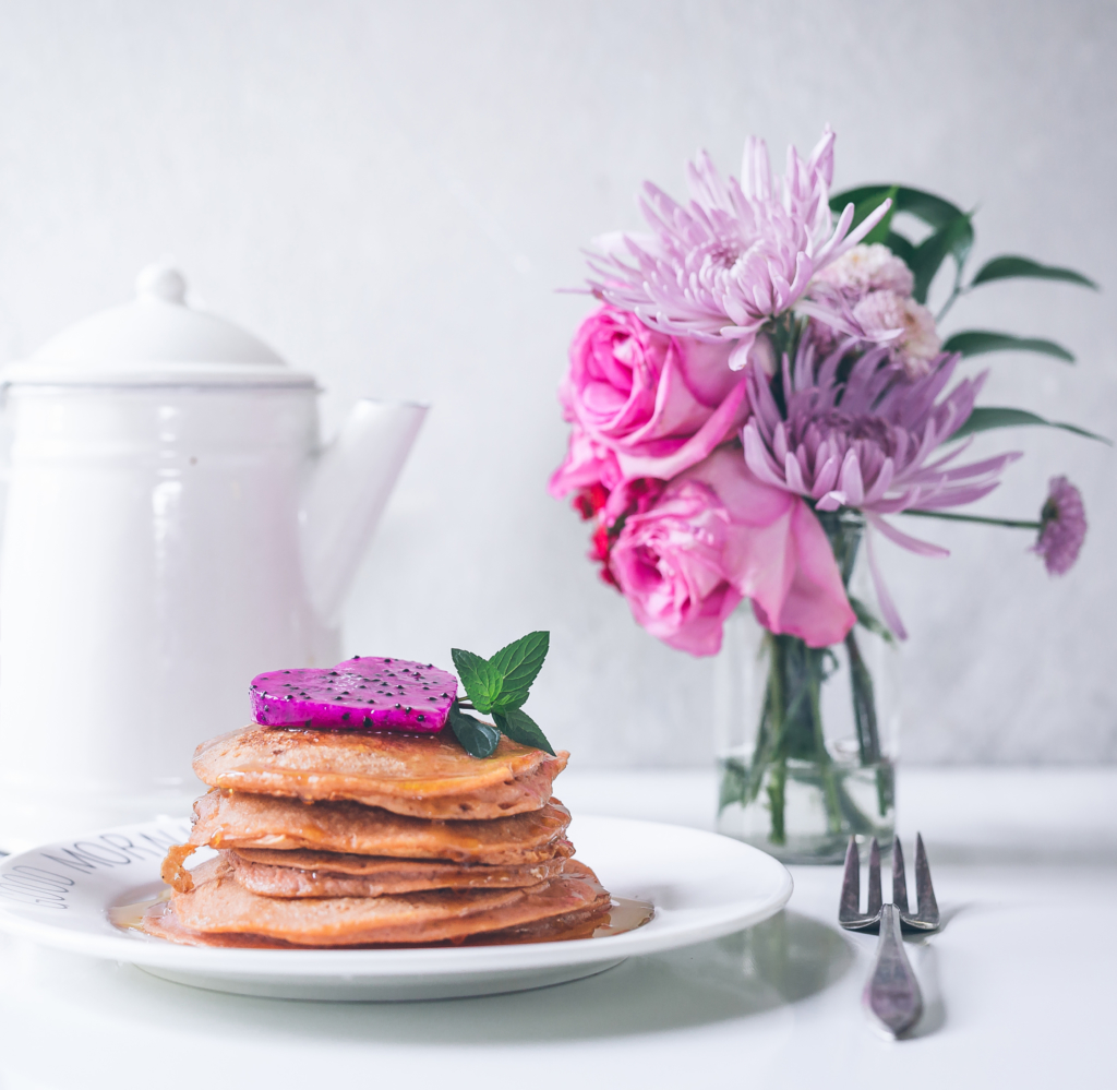 Stack of pancakes with a pink dragon fruit slice on top beside a bouquet and a teapot, elegant breakfast setup. Stack of pancakes with a pink dragon fruit slice on top beside a bouquet and a teapot, elegant breakfast setup.