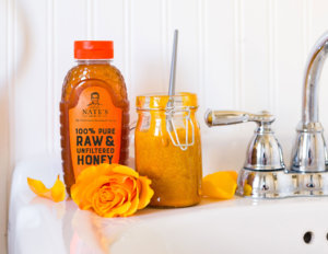 Nate's honey bottle next to jar of facial scrub with spoon on bathroom counter, orange flower decor.