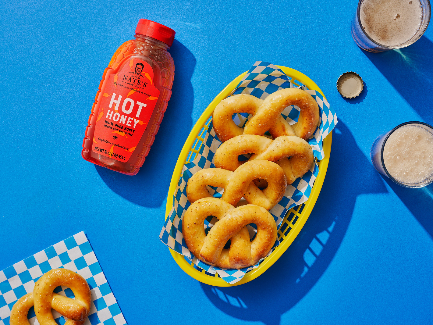  Nate’s hot honey bottle next to a basket of honeyed-pretzels on a blue background.