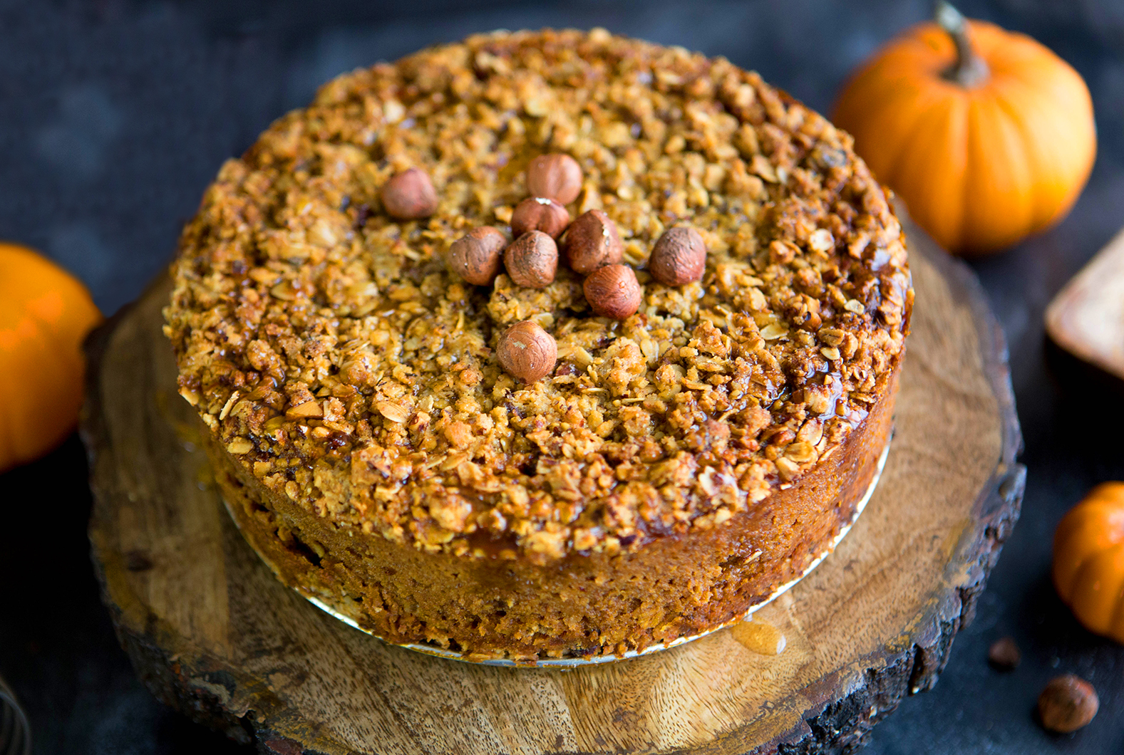 Hazelnut-topped pumpkin pie on a wooden round, surrounded by pumpkins on a dark surface.
