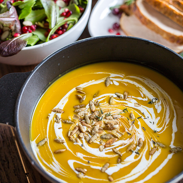 Creamy pumpkin and butternut soup in a pot, garnished with seeds and cream, with a salad in the background.