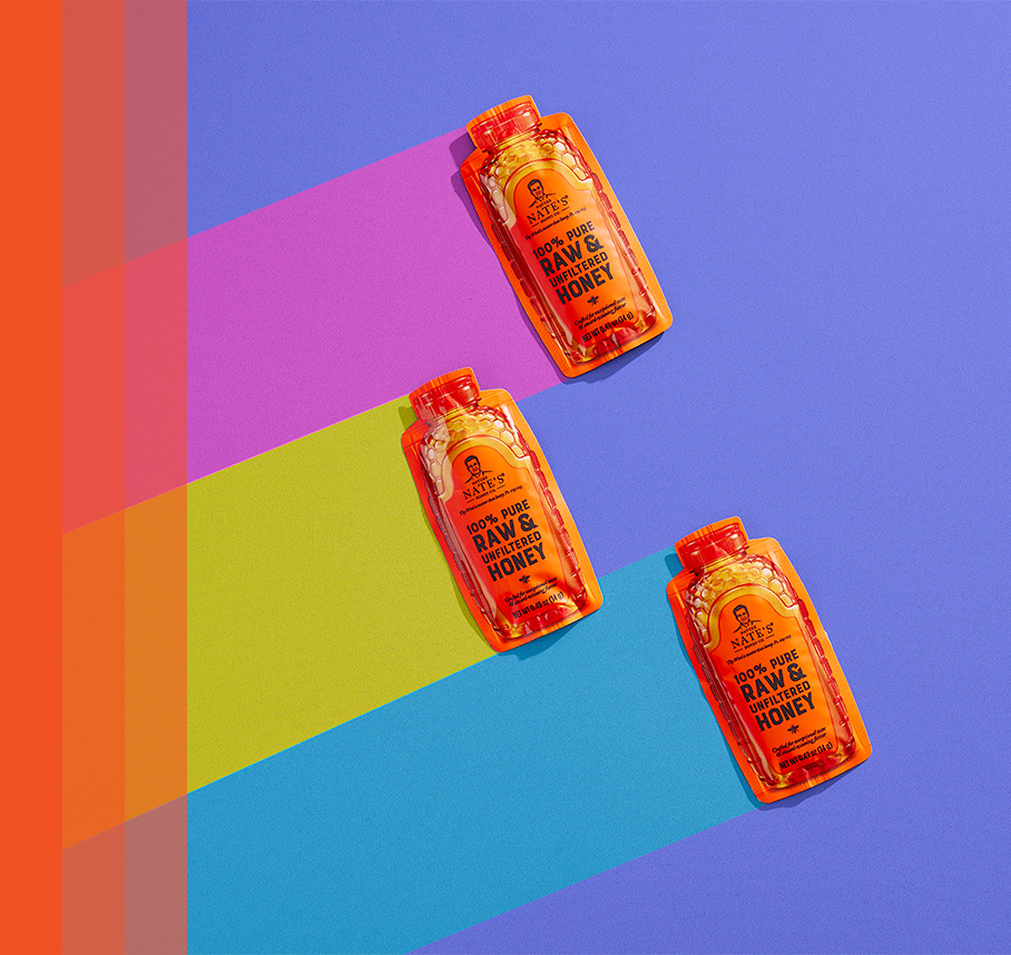 Three Nate's honey minis on a purple background with pink, lime green and blue streaming to the left of the packets.