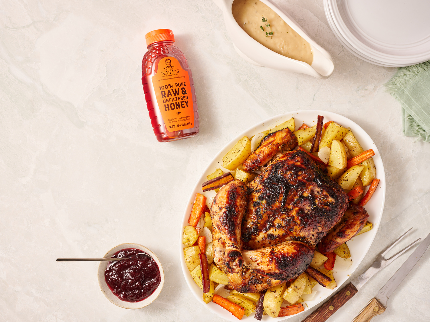Roasted chicken with vegetables, Nate’s honey bottle, cranberry sauce and gravy boat on a marbled surface.